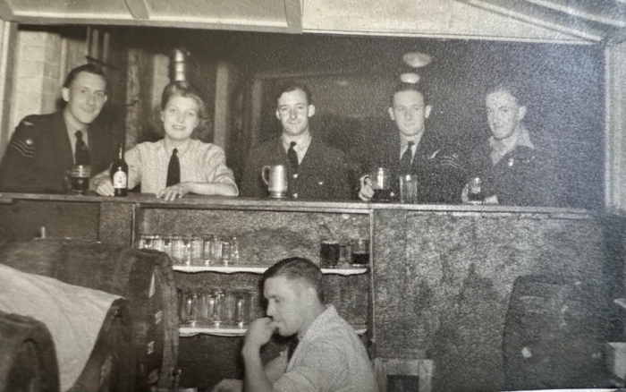 Black and white photo showing give air crew standing at a canteen with a sixth man pouring alcohol out of a barrel, 1940s.