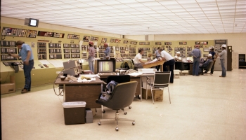 Colour photo of the interior of the control room at the AGE Plant in Joffre. Includes a bank of computers built into the wall, desks with chairs facing the wall in the middle of the room, and various men standing at the computer and desks. Room is very well-lit. 1979.