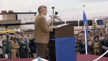 Colour photo of Premier Peter Lougheed standing at a blue and brown podium on a purple carpeted stage giving a speech for the opening of the AGE plant in Joffre, Alberta, in 1979. The audience is visible standing in the background.