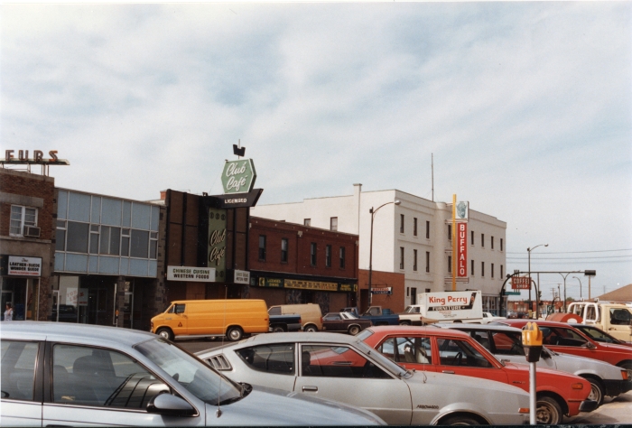 Colour photo of a streetscape with businesses including the Club Cafe and Buffalo Hotel across the road and parked cars in the the foreground. Red Deer, 1993.