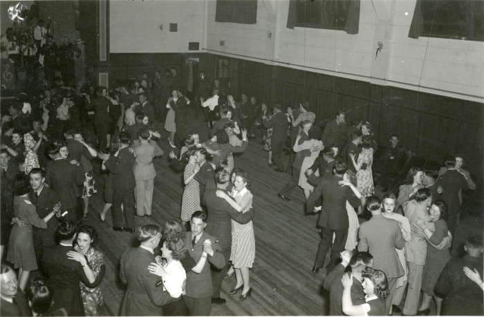 Black and white photo of a large crowd of young men in military uniform and women dancing inside large recreation hall at No. 36 Service Flying Training School