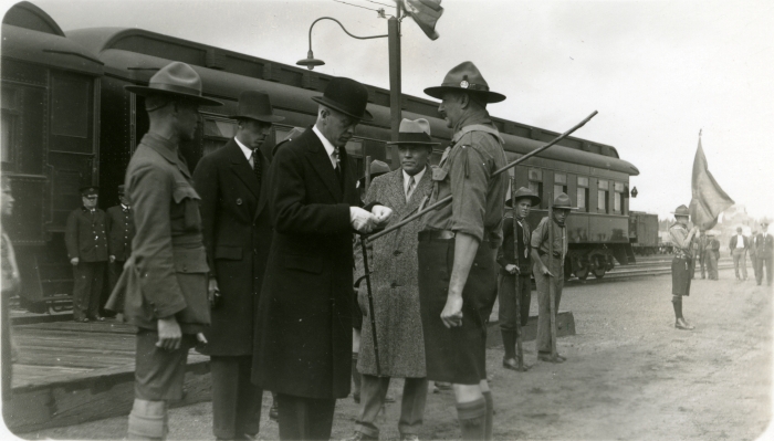 Black and white photo of a man presenting a medal to a man in a boy scout leader uniform in 1932. They are in front of a passenger train car and there are boy scouts as witnesses.