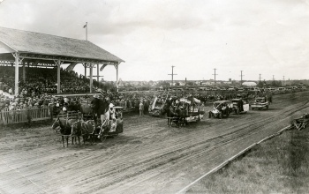 Black and white photo of parade floats passing the grandstands in the old Exhibition grounds
