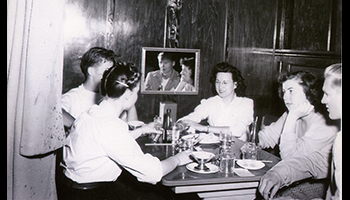 Black and white photo of a group of young adults at a booth style table in a restaurant in the 1950s