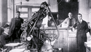 black and white photo of men standing around large printer machines, around 1916