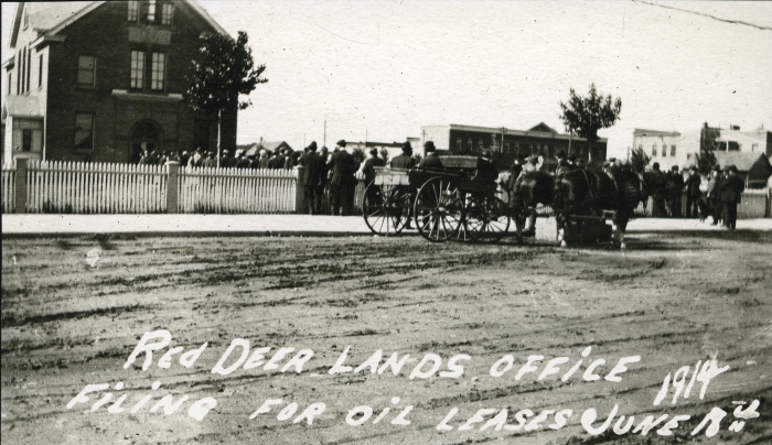 Black and white photo of crowds of people lining up outside the Red Deer Lands office in 1914. The building is a stone square and a dirt road is visible in the foreground.
