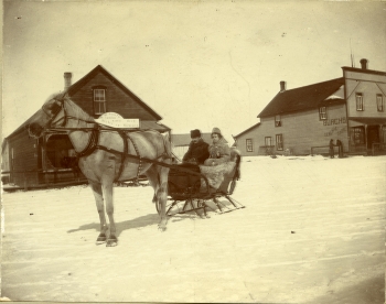 Black and white photo of R.C. Brumpton and his wife H. Drusilla Brumpton in cutter at Ross Street and Gaetz Avenue, with the NWMP barracks in the back