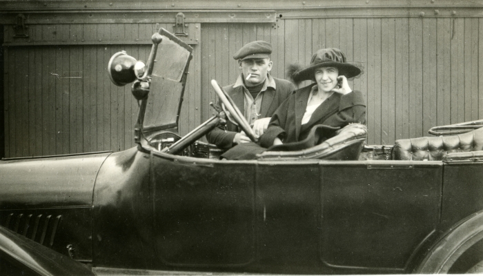 Black and white photo of a couple sitting in an open-toped car in the 1920s in Alberta. Both are wearing hats and coats.