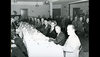 Black and white photo of a group of men in suits sitting at long tables with white table cloths in a banquet setting