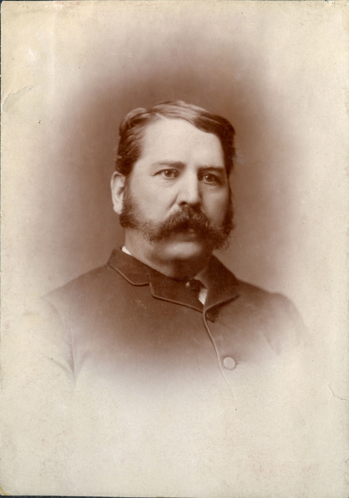 Sepia toned official portrait of a man with thick side burns and a large moustache. He is wearing a buttoned jacket.