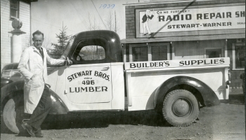 Black and white photo of a man standing next to a Stewart Brothers lumber struck outside a building labelled Trimble Radio Repair Shop