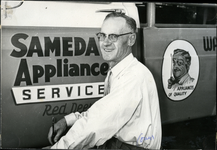 Black and white photo of a man smiling and facing the camera next to a hand-painted sign for Sameday Appliance Service