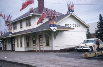 Coloured photo of the Canadian National Railway train station in Red Deer, Alberta, decorated with flags for the arrival of the last steam train